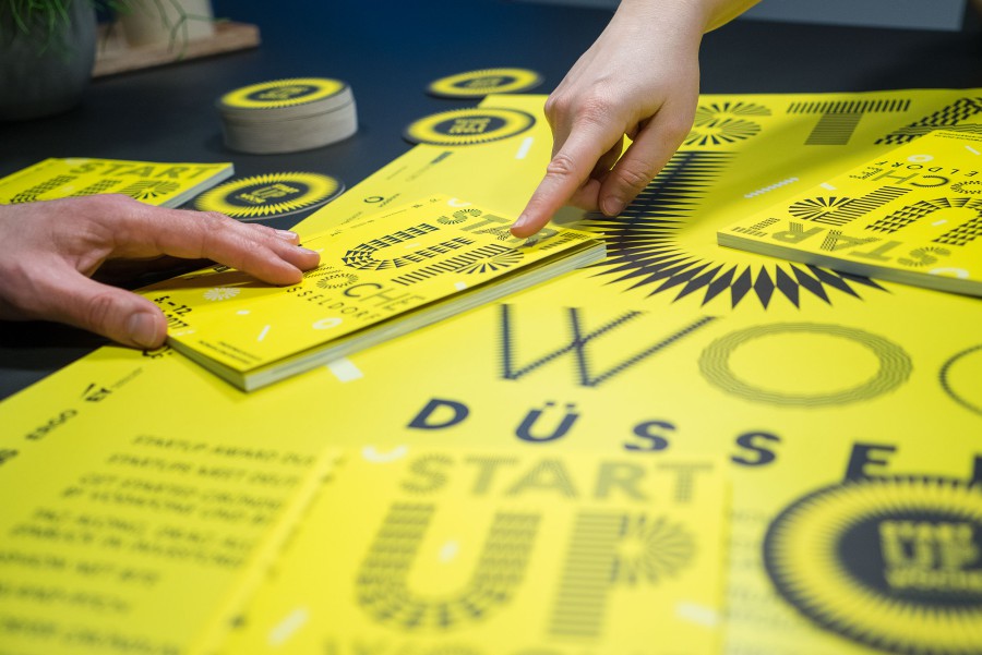 Hand points to the printed materials from Startup Week, including stickers, a program booklet, and posters