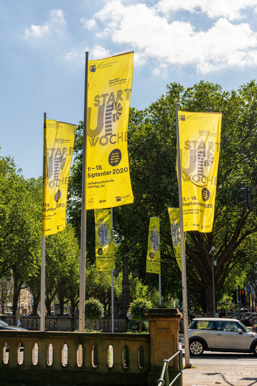 A row of yellow Startup Week flags flying from flagpoles in front of green trees along Düsseldorf's Kö