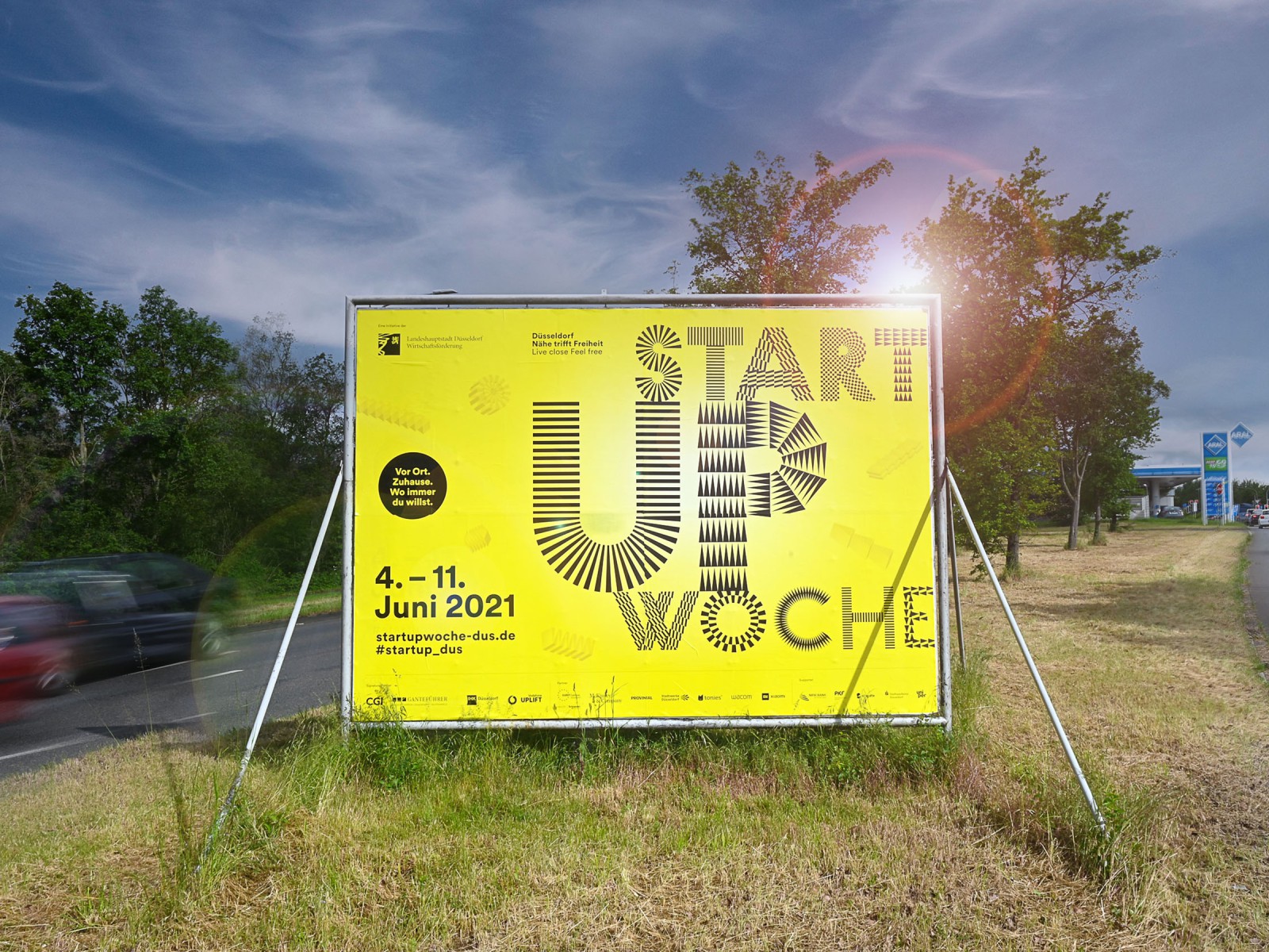 A freestanding large yellow poster for Düsseldorf Startup Week 2021 (June 4–11) on a traffic island, backlit by the evening sun and surrounded by trees and passing cars
