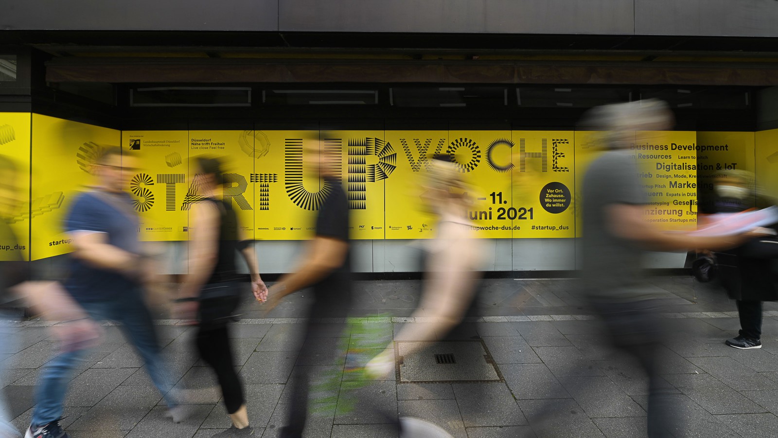 Wide-angle photo of downtown Düsseldorf featuring a large yellow “Startup Week” promotional banner, with blurry passersby walking by