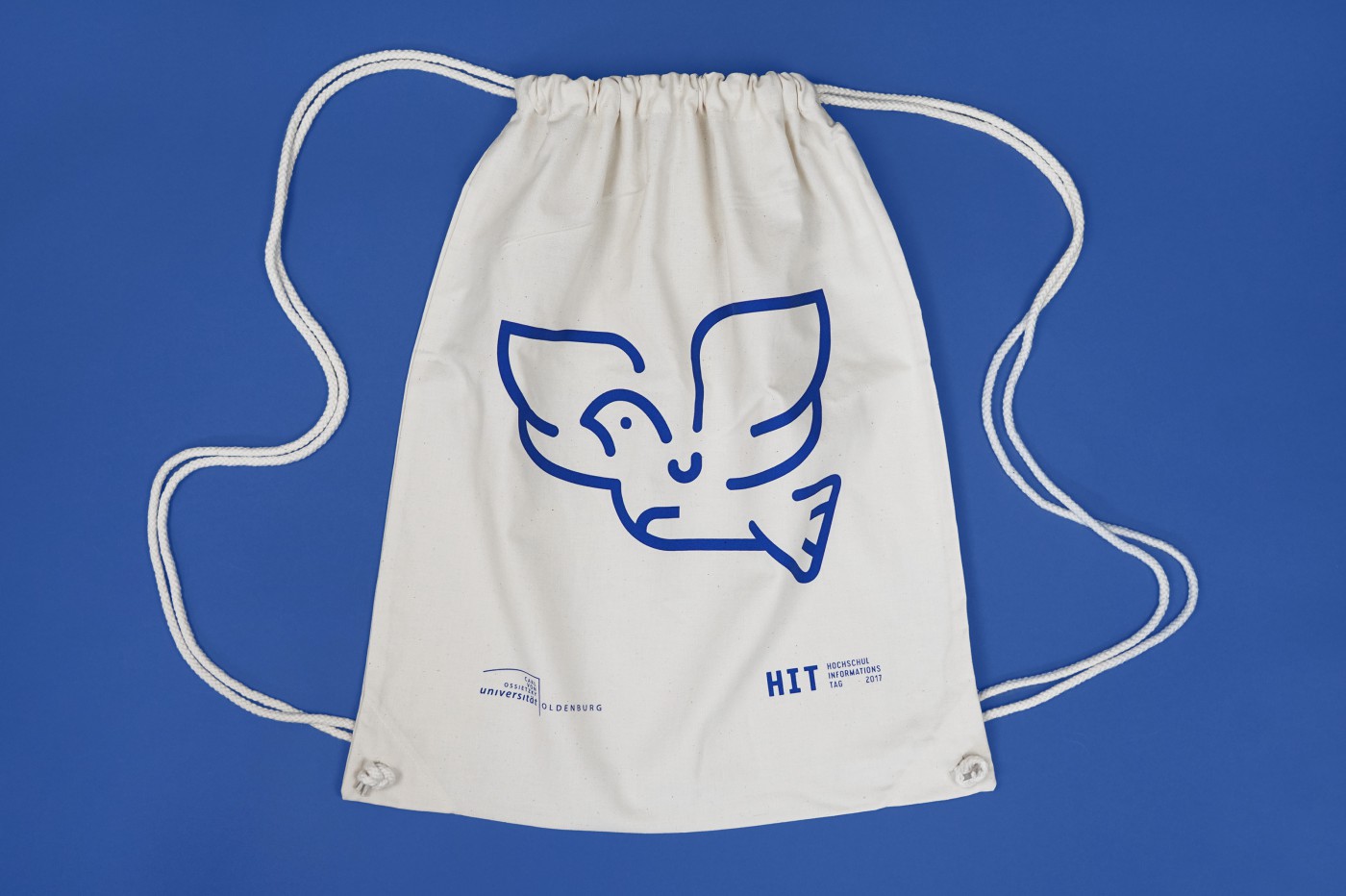 Heller Baumwoll-Turnbeutel mit blauer Linienzeichnung eines stilisierten fliegenden Vogels, Leuchtturms und Fahrrads sowie dem Logo der Carl von Ossietzky Universität Oldenburg und dem HIT-Schriftzug 2017 auf blauem Hintergrund